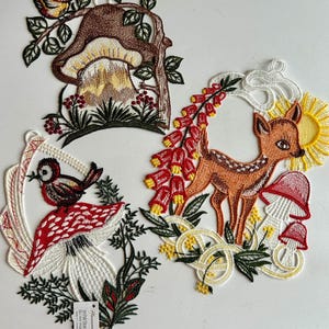 May include: Three embroidered felt ornaments featuring woodland creatures. The first ornament is a brown mushroom with a red-breasted bird perched on a branch. The second ornament is a red and white mushroom with a red-breasted bird perched on top. The third ornament is a fawn with a sun behind it and red flowers.