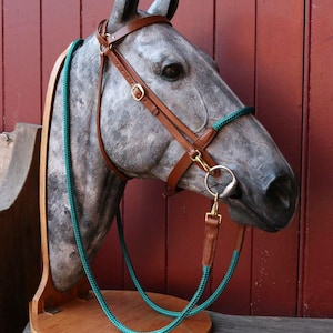 May include: A gray horse head sculpture with a brown leather bridle and a teal rope. The sculpture is mounted on a wooden base and set against a red wooden wall. The bridle has gold-colored metal hardware.