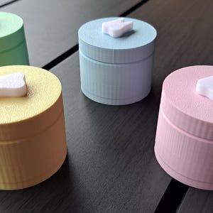 Könnte beinhalten: Vier kleine, runde, pastellfarbene Boxen mit einem weißen, zahnförmigen Deckel. Die Boxen sind in den Farbtönen Grün, Blau, Gelb und Rosa gehalten. Die Deckel sind strukturiert und die Boxen haben ein geripptes Design.