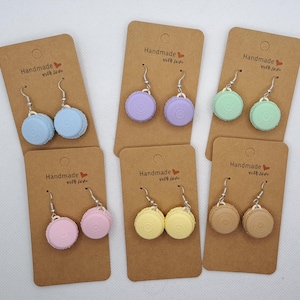 Könnte beinhalten: Ohrringe in Form von bunten Makronen in Pastelltönen: Blau, Lila, Grün, Rosa, Gelb und Braun. Jedes Paar wird auf einer braunen Karte mit dem Text "Handmade with love" präsentiert.