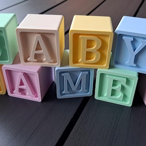 Peut inclure: Des blocs pour bébé aux couleurs pastel épelant "BABY NAME" et un cœur. Les blocs sont dans des tons de vert, jaune, rose, bleu et beige, avec les lettres et le cœur dans une couleur contrastante.