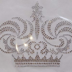 Könnte beinhalten: Ein Strass-Kronen-Design auf weißem Hintergrund. Die Krone zeigt silber- und goldfarbene Strasssteine, die in einem detaillierten Muster angeordnet sind. Das Kronendesign umfasst eine spitze Spitze und geschwungene Wirbel.