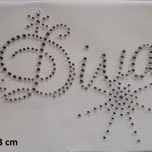 Könnte beinhalten: Ein weißer Stoffaufnäher mit den Maßen 10 x 13 cm, der das Wort Diva in Schreibschrift zeigt, geformt aus kleinen, klaren, reflektierenden Strasssteinen, akzentuiert durch zwei Sternmotive links und ein größeres Sternmuster auf der rechten Seite.