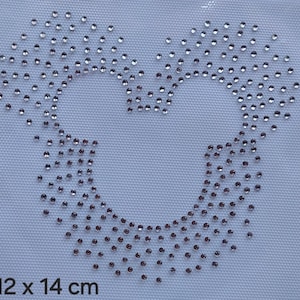 May include: A rhinestone applique in the shape of a heart, composed of numerous small, sparkling rhinestones. The applique is set against a white fabric background. The dimensions are 12 x 14 cm.