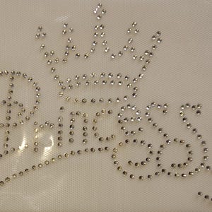 May include: A rhinestone design featuring the word "Princess" in a cursive font, topped with a crown. The rhinestones are clear and arranged on a white mesh background. The design is suitable for embellishing apparel or accessories.