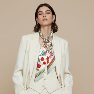 Op de afbeelding: Een crèmekleurige zijden sjaal met een bloemenprint van rode, roze en blauwe bloemen. De sjaal wordt gedragen door een persoon in een crèmekleurig pak. De sjaal heeft een lichtblauwe rand met gele, roze en groene strepen.