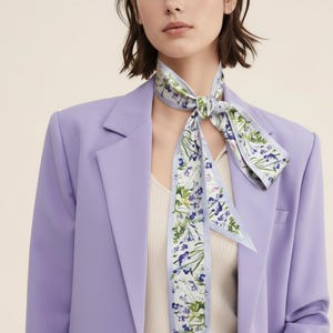 Könnte beinhalten: Hellblauer Schal mit floralem Muster, um den Hals gebunden. Der Schal zeigt blaue und grüne Blumen auf weißem Grund. Die Person trägt einen lila Blazer und ein cremefarbenes Oberteil.