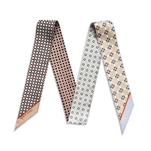 May include: Two silk scarves with geometric patterns. One is brown with white dots, the other is grey with a floral design, and the third is beige with a checkered pattern. The scarves are labelled "LUCY DALI" on the corner.