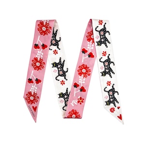 May include: A decorative scarf featuring a repeating pattern of black cat silhouettes and red flowers on a pink and white background. The scarf has the text "LUCY DALI" printed on it.