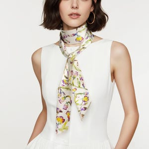 Peut inclure: Un foulard en soie crème avec un motif floral jaune, violet et vert. Le foulard est noué autour du cou, créant un accessoire élégant. Le motif présente des illustrations botaniques délicates.