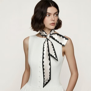 May include: A white silk scarf with a black scalloped edge is tied around the neck. The scarf has a small logo in the corner. The woman is wearing a white sleeveless dress.