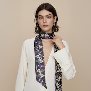 Könnte beinhalten: Gemusterter Seidenschal mit dunklem Hintergrund und geometrischen und floralen Designs in Lila-, Weiß- und Goldtönen. Der Schal wird um den Hals getragen und fällt über die Vorderseite einer weißen Bluse.