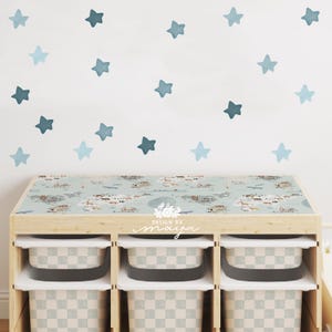May include: A wooden storage unit with three cubbies, each holding a checkered bin. The top surface features a world map design. Blue star decals are on the white wall above the unit. The text "DESIGN BY maya" is on the front.