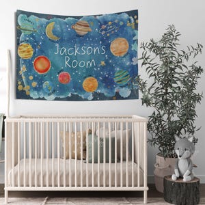 Pode incluir: Uma tape&ccedil;aria em estilo aquarela com uma cena espacial azul com planetas, estrelas e o texto "Jacksons Room". Os planetas s&atilde;o em v&aacute;rias cores, incluindo amarelo, laranja e azul. A tape&ccedil;aria est&aacute; pendurada acima de um ber&ccedil;o branco.