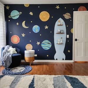 May include: A dark blue wall with a space-themed mural featuring planets, stars, and a rocket ship. A white armchair with a blue throw blanket sits in front of the wall. A small wooden side table with three drawers is next to the chair. A blue and white rug is on the floor in front of the chair.