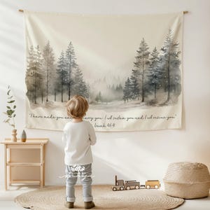 May include: A beige wall tapestry with a watercolor forest scene in shades of gray and blue. The text "I have made you and carry you...Isaiah 46:4" is printed on the bottom. A child stands in front of the tapestry.