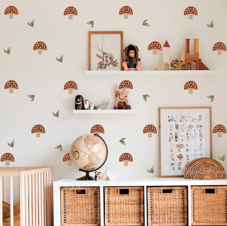 K&ouml;nnte beinhalten: Eine wei&szlig;e Wand mit braunen Pilz- und gr&uuml;nen Blatt-Wandaufklebern. Die Wand ist mit einem Globus, einem Holzregal mit Spielzeug und einer wei&szlig;en Kommode mit Korb-Schubladen dekoriert.