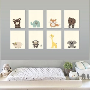 May include: A white changing table with a white changing pad with black polka dots. The changing table has three drawers with animal-shaped drawer pulls. Above the changing table is a gray wall with eight framed prints of different animals. The animals include a bear, elephant, fox, lion, sheep, monkey, giraffe, and koala.