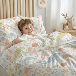 May include: A white bedding set with a colorful ocean-themed pattern featuring seahorses, jellyfish, starfish, and a whale. A young child is lying in the bed, smiling.