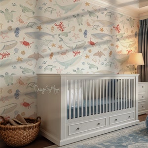 May include: A white crib with two drawers, set against a wall with a sea life pattern. The wallpaper features whales, turtles, starfish, and other sea creatures in soft colors. A woven basket and a blue patterned rug complete the nursery setting.