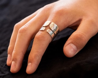 May include: A silver-toned ring with a textured, hammered design. The ring features an open design with two overlapping sections, creating a unique and modern look. The ring is worn on a finger, showcasing its intricate details and craftsmanship.