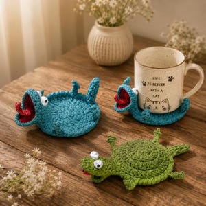 Può includere: Due sottobicchieri rotondi all'uncinetto a forma di creature con occhi mobili, uno verde acqua e uno verde, con bocche aperte e lingue rosse. Una tazza in ceramica con il testo Life is better with a cat e un muso di gatto poggia sul sottobicchiere.