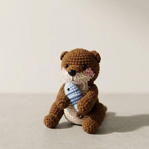 Könnte beinhalten: Ein braunes gehäkeltes Otter-Stofftier mit cremefarbenem Bauch, rosa gestickten Wangen, schwarzen Perlenaugen, einer schwarzen Nase und weißen Faden-Schnurrhaaren. Der Otter hält einen kleinen, hellblauen gehäkelten Fisch zwischen seinen Vorderpfoten.