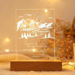 May include: A rectangular clear acrylic plaque with a white etched mountain landscape, a sun, and the text I can do all this through him who gives me strength Philippians 4:13, mounted on a rectangular wooden base with an integrated light.