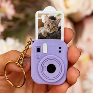 Puede incluir: Un llavero con forma de cámara de color lavanda con una cadena y un llavero dorados. La cámara tiene una foto de un gato. La cámara tiene un flash, una lente y dos pequeños puntos negros. El gato de la foto es gris y marrón.