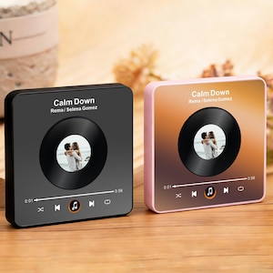 May include: Two square music player plaques featuring a black vinyl record graphic with a photo of a couple in the centre. The text reads Calm Down by Rema and Selena Gomez, with playback controls, a progress bar, and timestamps 0:01 and 3:56.