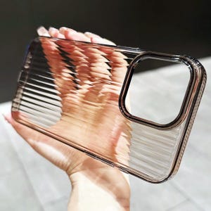 Puede incluir: Funda transparente para teléfono con diseño acanalado. La funda tiene un borde metálico alrededor de la abertura de la cámara. La funda se sostiene en una mano.