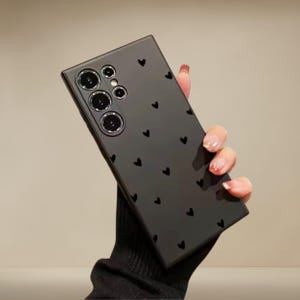 May include: A black phone case with a pattern of small black hearts. The case has a glossy finish and features a camera array on the upper left corner. The phone case is held by a person wearing a black sleeve.