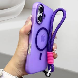 Puede incluir: Una funda de teléfono morada con un cordón a juego. La funda tiene un diseño circular con un logotipo de Apple y una pequeña línea negra. El cordón tiene un acento rosa y un cierre blanco.