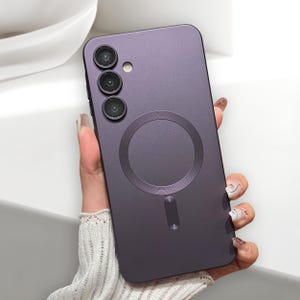 Op de afbeelding: Een donkerpaarse telefoonhoes met een cirkelvormige magnetische bevestiging en drie cameralenzen. De hoes wordt vastgehouden door een hand met verzorgde nagels, die het ontwerp en de functies van de telefoon benadrukt.