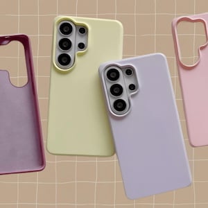 May include: A collection of colorful phone cases in shades of yellow, lavender, and pink. The cases are designed to fit a phone with a multi-lens camera system. The cases are displayed on a beige grid background.