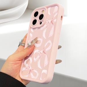 Pode incluir: Capa de telefone rosa claro com um padrão repetido de laços brancos. A capa tem uma borda elevada ao redor da câmera e um acabamento liso. Projetada para um smartphone.