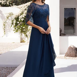 Op de afbeelding: Een marineblauwe avondjurk met een doorschijnend, versierd lijfje en een zwierige rok. De jurk heeft een delicate overlay op de schouders en een getailleerde taille. Het model draagt zwarte hakken.