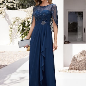 Op de afbeelding: Een marineblauwe formele jurk met een doorschijnend, versierd lijfje en een zwierige rok. De jurk heeft een cape-achtig mouwontwerp en een versierde riem. Een zilveren clutch maakt de look compleet. Geschikt voor speciale gelegenheden.