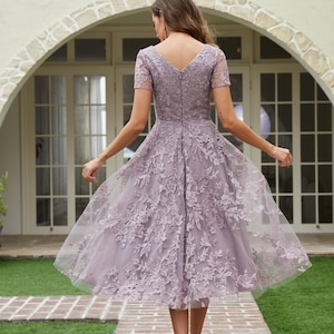 Peut inclure: Robe lavande mi-longue avec un col en V et des manches courtes. La robe pr&eacute;sente des d&eacute;tails de dentelle florale et une jupe fluide. Le mod&egrave;le porte des talons argent&eacute;s et marche sur un chemin de briques.