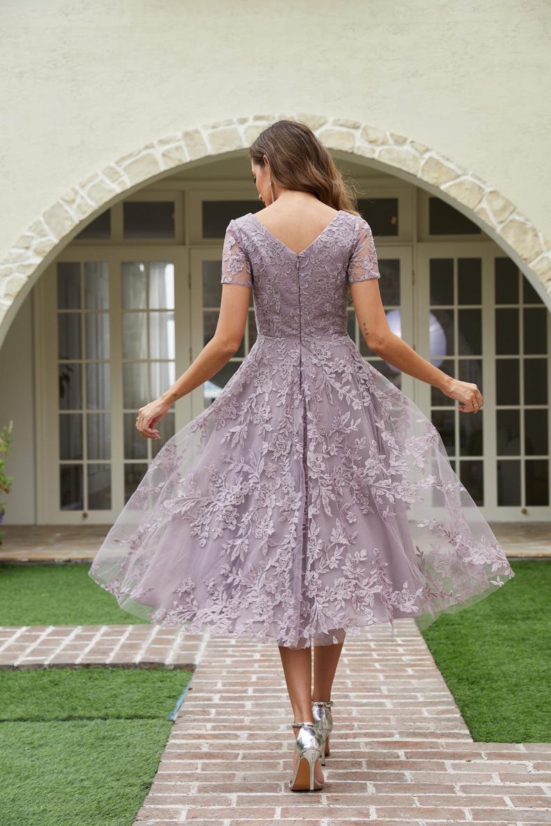 Peut inclure: Robe lavande mi-longue avec un col en V et des manches courtes. La robe pr&eacute;sente des d&eacute;tails de dentelle florale et une jupe fluide. Le mod&egrave;le porte des talons argent&eacute;s et marche sur un chemin de briques.