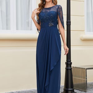 Op de afbeelding: Een marineblauwe avondjurk met een doorschijnend, versierd lijfje en een zwierige rok. De jurk heeft een delicate cape-achtige mouwdetail en een versierde riem. Het elegante ontwerp is geschikt voor speciale gelegenheden.