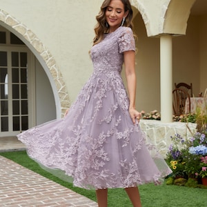 Peut inclure: Une robe lavande mi-longue avec des d&eacute;tails de dentelle florale. La robe a des manches courtes et une jupe fluide. Le mod&egrave;le porte des talons hauts argent&eacute;s. L'arri&egrave;re-plan comprend une all&eacute;e en briques et un b&acirc;timent avec des portes cintr&eacute;es.