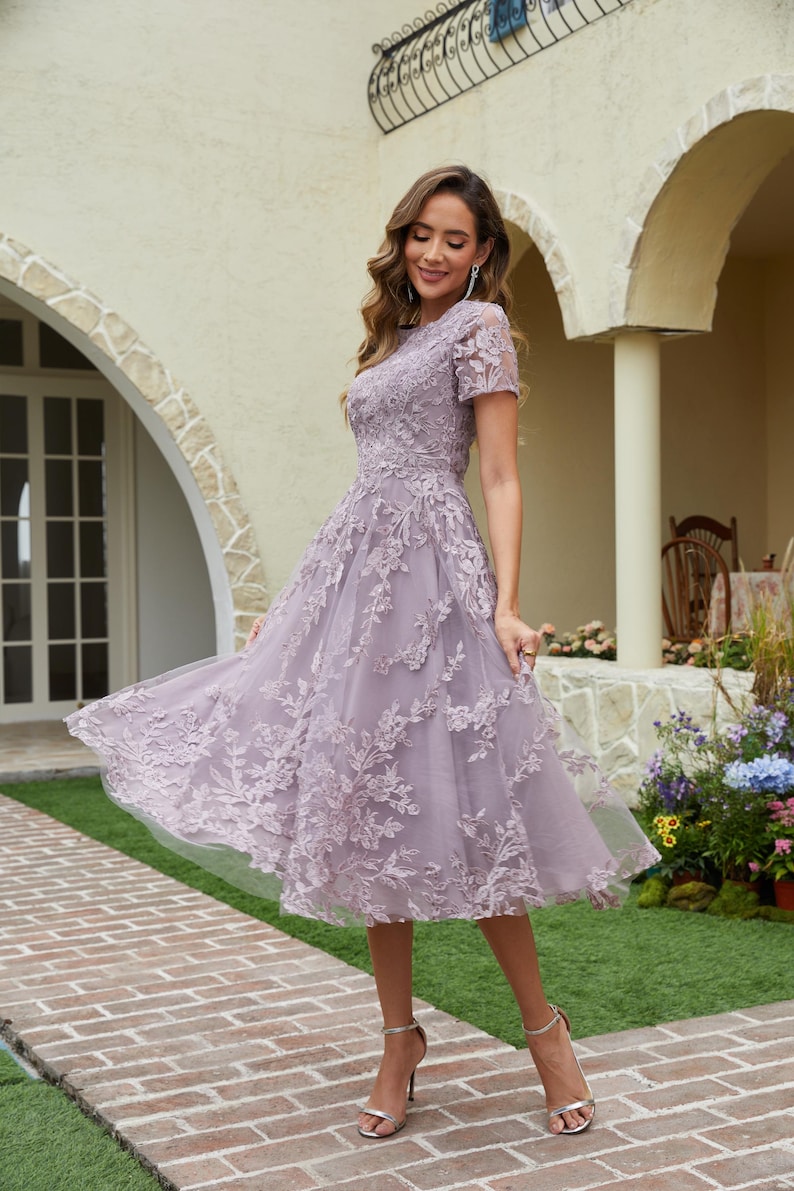 Peut inclure: Une robe lavande mi-longue avec des d&eacute;tails de dentelle florale. La robe a des manches courtes et une jupe fluide. Le mod&egrave;le porte des talons hauts argent&eacute;s. L'arri&egrave;re-plan comprend une all&eacute;e en briques et un b&acirc;timent avec des portes cintr&eacute;es.