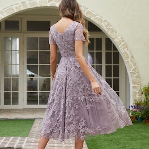 Peut inclure: Robe lilas mi-longue avec un col en V et des manches courtes. La robe pr&eacute;sente des d&eacute;tails de dentelle florale et une jupe fluide. La personne porte des talons hauts argent&eacute;s. L'arri&egrave;re-plan comprend une all&eacute;e en briques et un b&acirc;timent avec une porte cintr&eacute;e.
