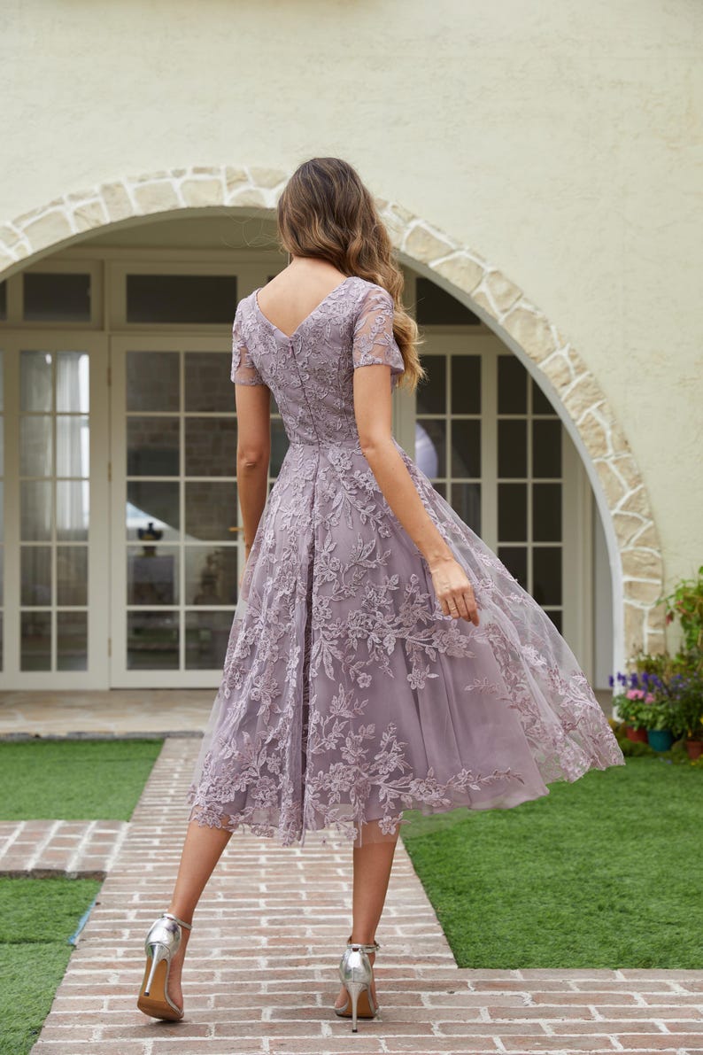 Peut inclure: Robe lilas mi-longue avec un col en V et des manches courtes. La robe pr&eacute;sente des d&eacute;tails de dentelle florale et une jupe fluide. La personne porte des talons hauts argent&eacute;s. L'arri&egrave;re-plan comprend une all&eacute;e en briques et un b&acirc;timent avec une porte cintr&eacute;e.