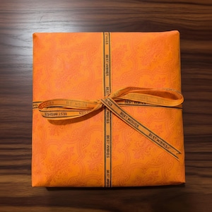 Op de afbeelding: Een oranje geschenkdoos verpakt in papier met patronen en vastgebonden met een lint. Op het lint staat de tekst "BEST WISHES". De doos staat op een houten oppervlak.