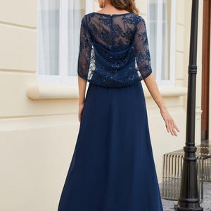 Op de afbeelding: Een marineblauwe formele jurk met een doorschijnende, versierde overlay. De overlay heeft ingewikkelde bloemenborduursels en sprankelende accenten. De lange, vloeiende rok is gemaakt van een gladde, effen stof. De jurk is geschikt voor speciale gelegenheden.