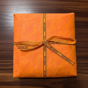 Peut inclure: Un coffret cadeau orange envelopp&eacute; dans du papier &agrave; motifs, nou&eacute; avec un ruban portant l'inscription "BEST WISHES". La bo&icirc;te est pos&eacute;e sur une surface en bois. Le papier d'emballage a un motif floral.