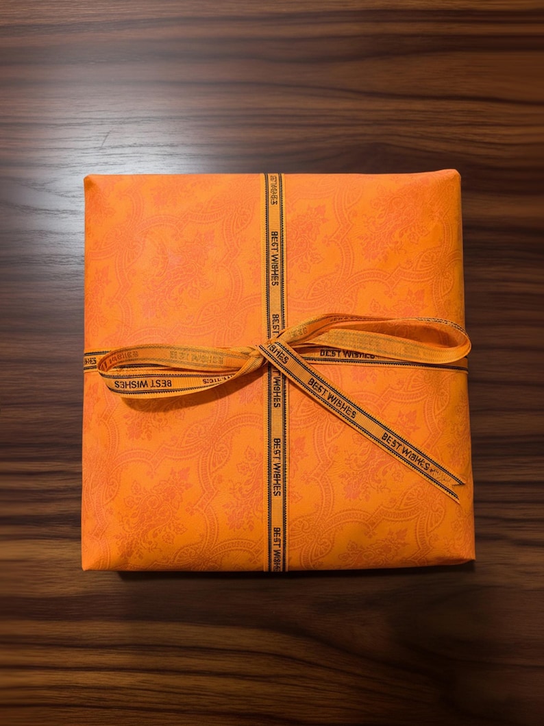 Peut inclure: Un coffret cadeau orange envelopp&eacute; dans du papier &agrave; motifs, nou&eacute; avec un ruban portant l'inscription "BEST WISHES". La bo&icirc;te est pos&eacute;e sur une surface en bois. Le papier d'emballage a un motif floral.