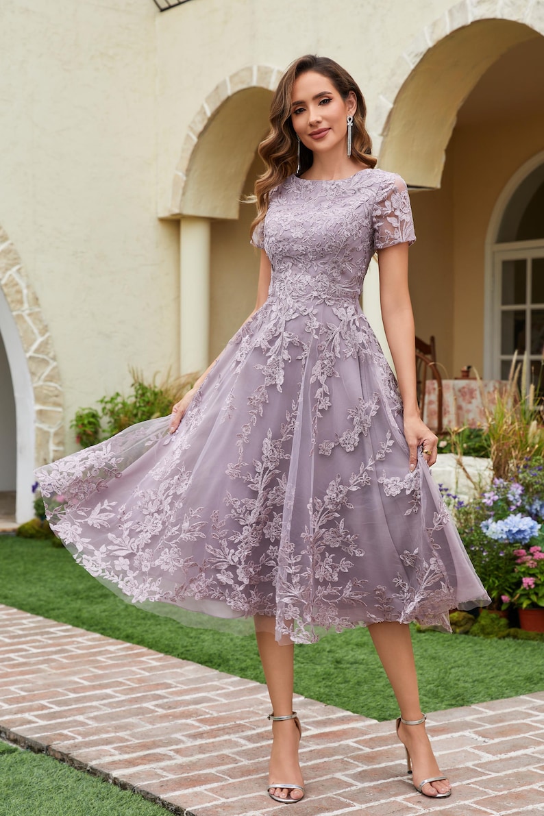 Peut inclure: Robe lavande, longueur genou, manches courtes. La robe pr&eacute;sente des d&eacute;tails de dentelle florale complexes sur un tissu transparent. La mod&egrave;le porte des talons argent&eacute;s. Convient aux &eacute;v&eacute;nements formels.